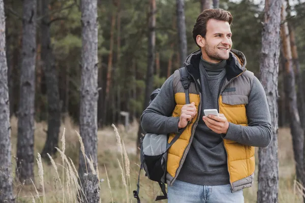 Young and happy man with smartphone and backpack hiking in forest — Stock Photo