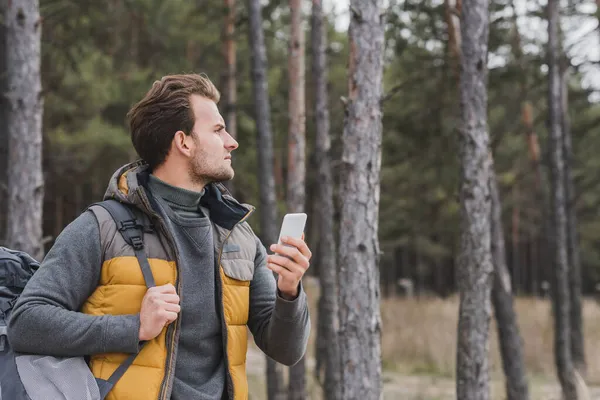 Homem com mochila e telefone celular olhando para longe na floresta de outono — Fotografia de Stock