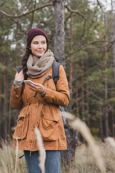 Smiling woman with smartphone looking away while hiking in autumn forest — Stock Photo