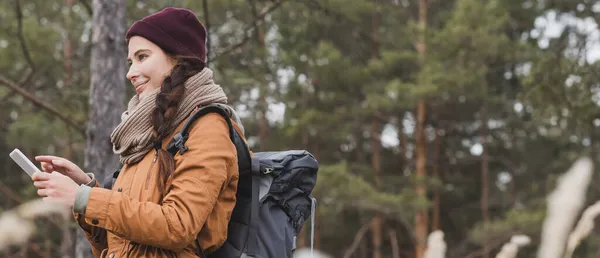 Mujer sonriente apuntando al teléfono inteligente mientras busca dirección durante el senderismo en el bosque, pancarta - foto de stock