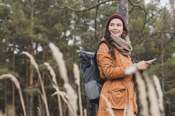 Улыбающаяся женщина с сотовым телефоном и рюкзаком смотрит в сторону во время похода на размытый передний план — стоковое фото