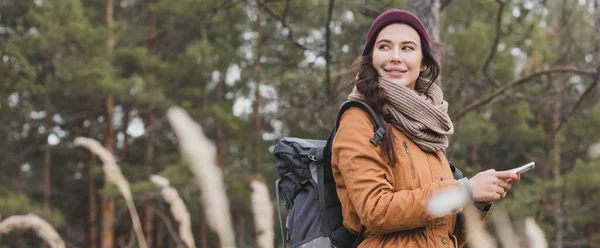 Mulher positiva com mochila e telefone celular caminhadas na floresta em primeiro plano turvo, banner — Fotografia de Stock