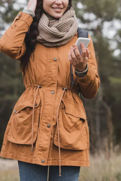 Vista recortada de la mujer sonriente en ropa de otoño sosteniendo el teléfono móvil durante el paseo en el bosque - foto de stock