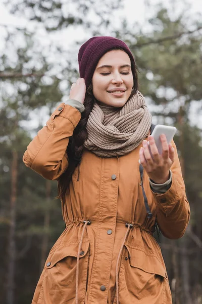 Cheerful young woman looking at mobile phone while walking in autumn forest — Stock Photo