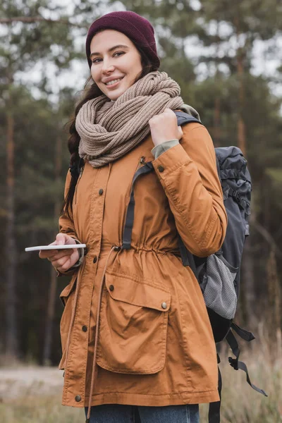 Jovem positiva com mochila e celular olhando para a câmera enquanto caminhava na floresta — Fotografia de Stock