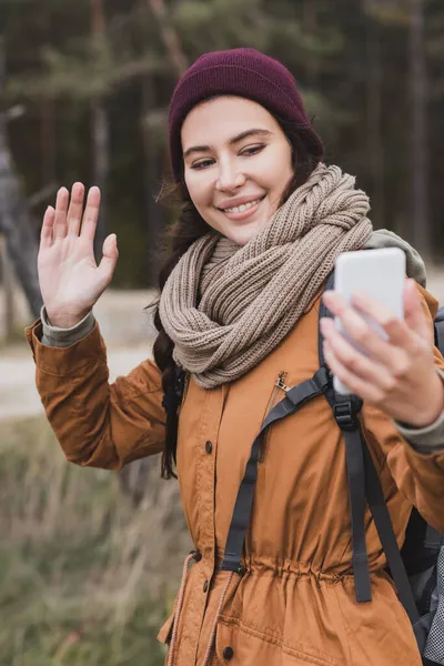 Mulher alegre na roupa de outono acenando mão durante chamada de vídeo em smartphone borrado na floresta — Fotografia de Stock