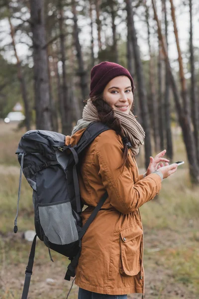 Allegra giovane donna sorridente alla macchina fotografica mentre cammina nella foresta con zaino e telefono cellulare — Foto stock