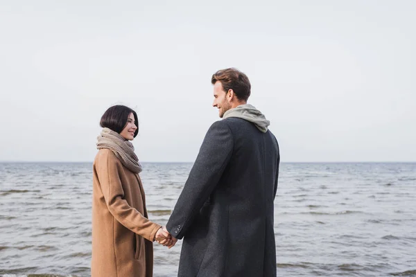 Young and happy couple in autumn clothes holding hands and looking at each other near sea — Stock Photo