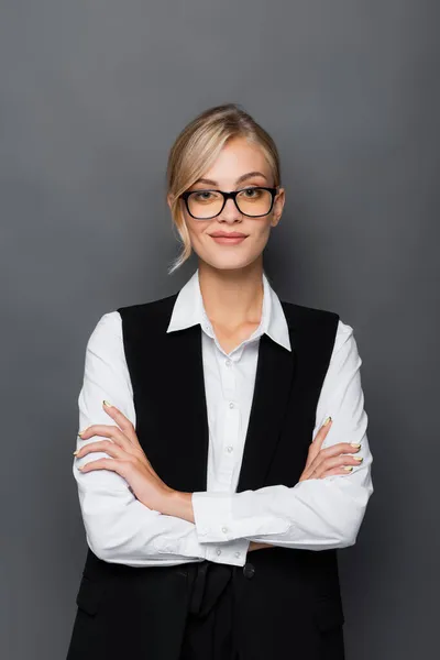 Lächelnde Geschäftsfrau mit Brille steht mit verschränkten Armen isoliert auf grau — Stockfoto