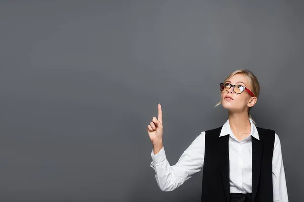 Blonde businesswoman in eyeglasses pointing with finger and looking up isolated on grey — Stock Photo