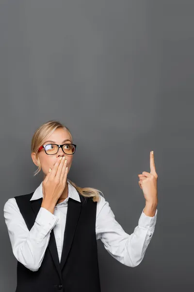Shocked businesswoman covering mouth and pointing with finger isolated on grey — Stock Photo
