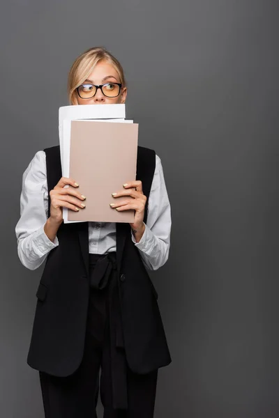 Blonde businesswoman in eyeglasses holding paper folders near face isolated on grey — Stock Photo