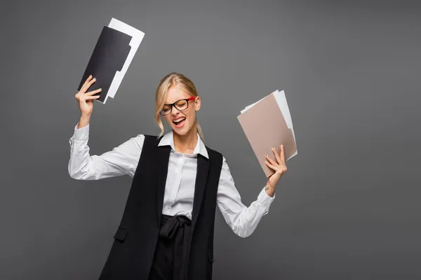 Fröhliche Geschäftsfrau in Weste und Brille hält Papiermappen isoliert auf grau — Stockfoto