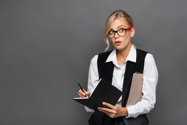 Junge Geschäftsfrau mit Brille schreibt auf Notizbuch und hält Papiermappen isoliert auf grau — Stockfoto