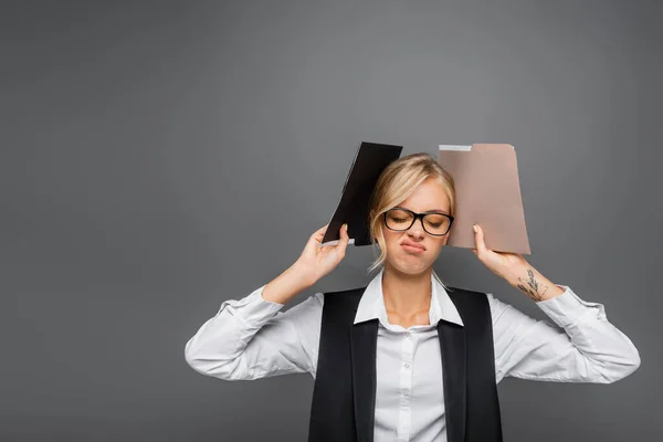 Exhausted businesswoman in eyeglasses holding paper folders isolated on grey — Stock Photo