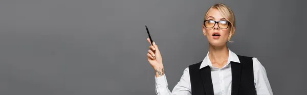 Pensive бізнес-леді в окулярах вказуючи рукою, тримаючи ручку ізольовано на сірому, банер — стокове фото