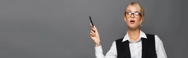 Pensive businesswoman in eyeglasses pointing with hand while holding pen isolated on grey, banner — Stock Photo