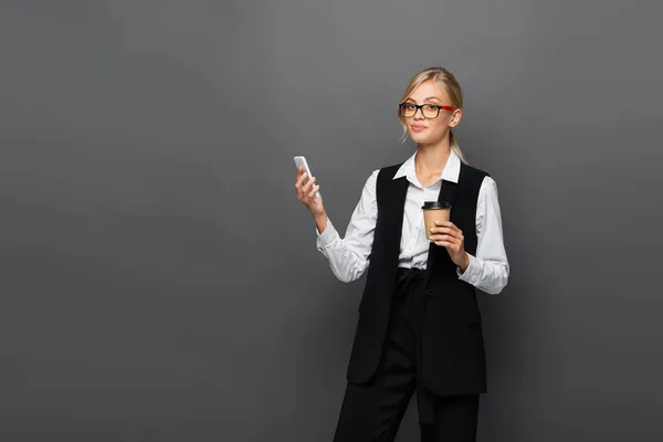 Empresária sorridente segurando smartphone e café para ir isolado em cinza — Fotografia de Stock