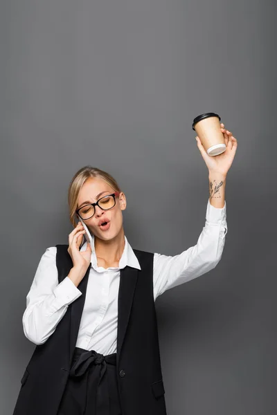 Захоплена бізнес-леді тримає паперовий стаканчик і розмовляє на смартфоні ізольовано на сірому — стокове фото