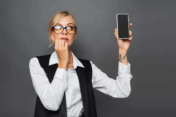 Mujer de negocios pensativa sosteniendo teléfono inteligente con pantalla en blanco aislado en gris - foto de stock