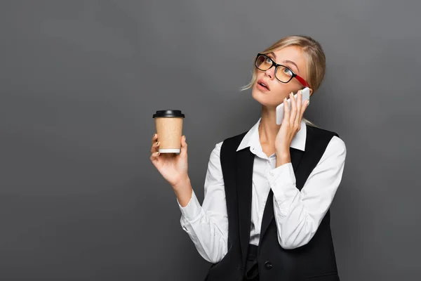 Pensive блондинка бізнесмен розмовляє по смартфону і тримає каву, щоб ізолювати на сірому — стокове фото