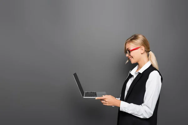 Vista lateral de la mujer de negocios sonriente en chaleco usando el ordenador portátil sobre fondo gris - foto de stock