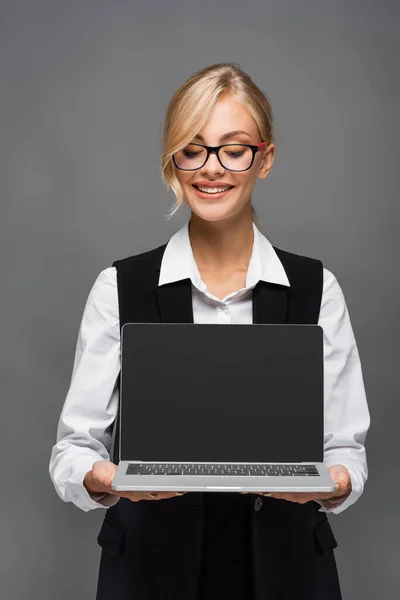 Mujer de negocios rubia en gafas que sostiene el ordenador portátil con pantalla en blanco aislado en gris - foto de stock