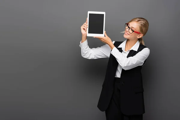 Mujer de negocios sonriente en gafas que sostiene la tableta digital con la pantalla en blanco en gris - foto de stock