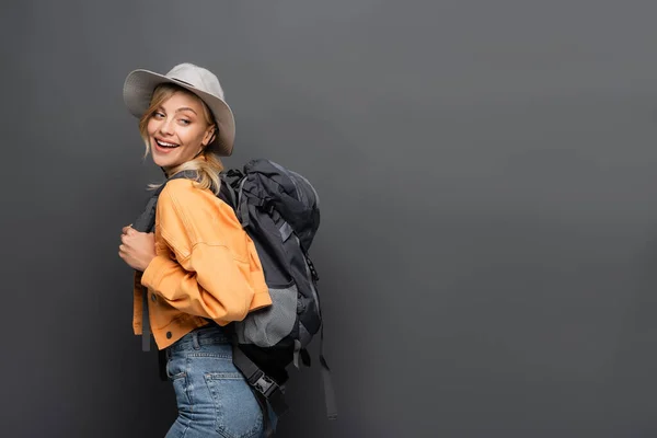 Turista positivo con mochila y sombrero mirando hacia otro lado aislado en gris - foto de stock