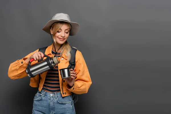 Lächelnder Tourist mit Rucksack, Thermoskanne und Tasse isoliert auf grau — Stockfoto