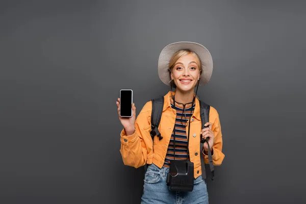 Улыбающийся блондин-турист со смартфоном и винтажной камерой, изолированной на сером — стоковое фото