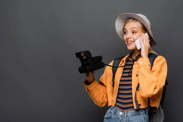Turista rubia en sombrero hablando en smartphone y sosteniendo cámara vintage aislada en gris - foto de stock