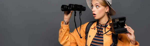 Amazed traveler holding vintage camera and looking at binoculars isolated on grey, banner — Stock Photo