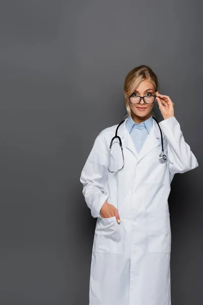 Молодий лікар тримає окуляри і дивиться на камеру ізольовано на сірому — стокове фото