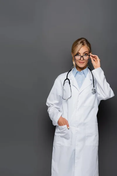 Joven médico sosteniendo anteojos y mirando a la cámara aislada en gris - foto de stock