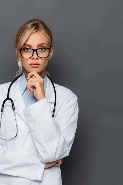 Pensive doctor in white coat and eyeglasses looking at camera isolated on grey — Stock Photo