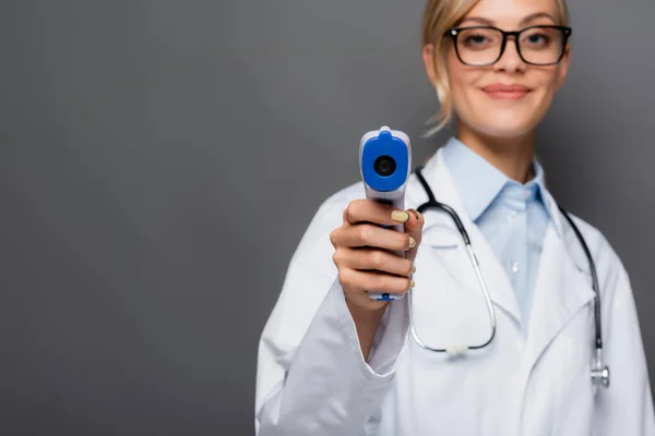 Pirómetro en la mano del médico borroso mirando a la cámara aislada en gris - foto de stock