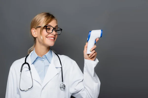 Fröhlicher Arzt schaut auf kontaktloses Pyrometer isoliert auf grau — Stockfoto