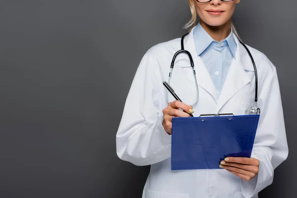 Vue recadrée du jeune médecin écrivant sur presse-papiers isolé sur gris — Photo de stock