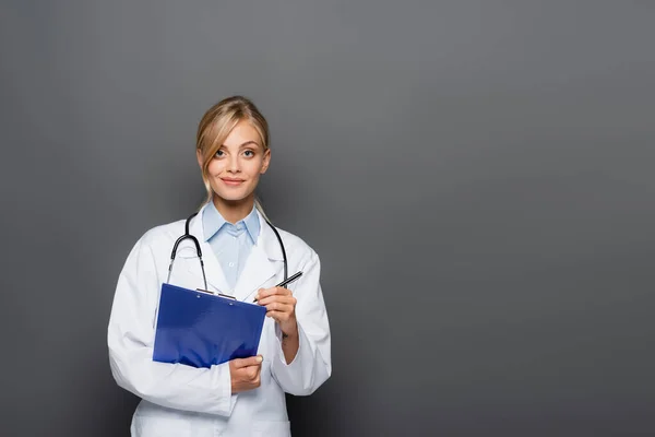 Blonde doctor holding pen and clipboard while looking at camera isolated on grey — Stock Photo