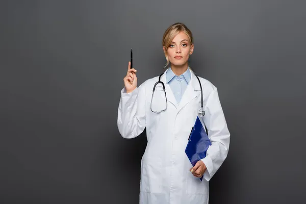 Médico loiro segurando prancheta e apontando com a mão sobre fundo cinza — Fotografia de Stock