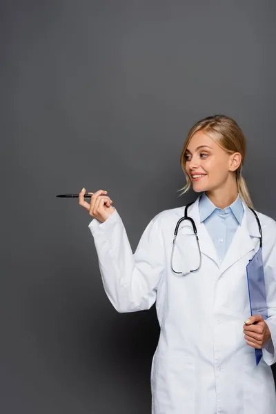 Lächelnder Arzt mit Klemmbrett, das mit der Hand auf grauem Hintergrund zeigt — Stockfoto