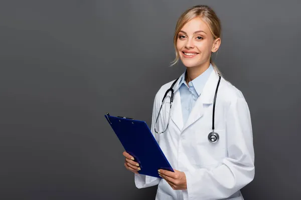 Happy young doctor holding clipboard and looking at camera isolated on grey — Stock Photo