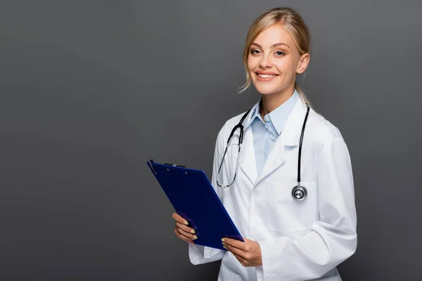 Feliz joven médico sujetando el portapapeles y mirando a la cámara aislada en gris - foto de stock