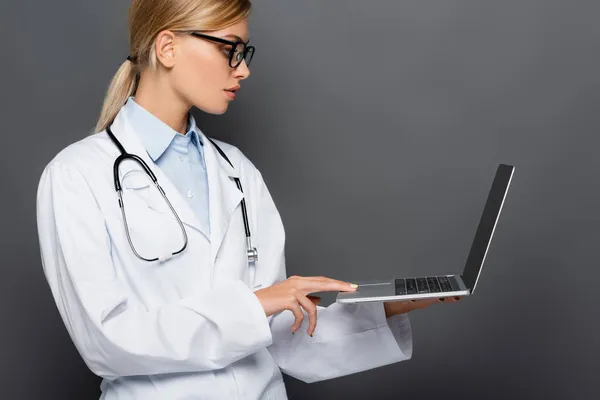 Side view of doctor in eyeglasses using laptop isolated on grey — Stock Photo