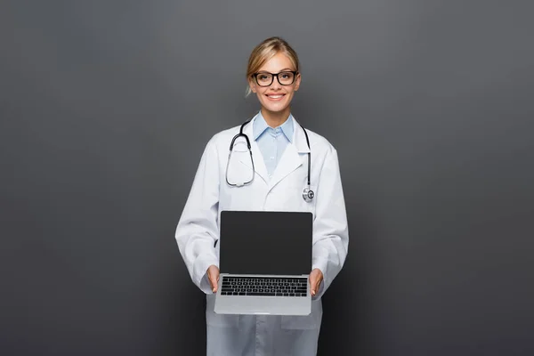 Позитивный доктор в белом халате показывает ноутбук с чистым экраном, изолированным на сером — стоковое фото