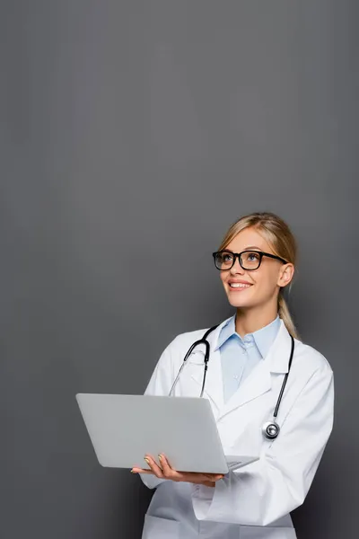 Médico sonriente en gafas con portátil y mirando hacia otro lado aislado en gris - foto de stock