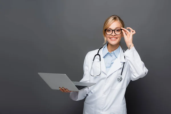 Feliz médico sosteniendo las gafas y el ordenador portátil mientras mira la cámara aislada en gris - foto de stock