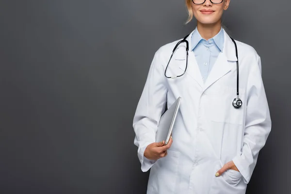 Обрізаний вид усміхненого лікаря, який тримає ноутбук і руку в кишені білого пальто ізольовано на сірому — стокове фото
