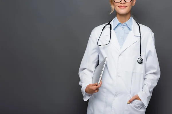 Vista recortada del médico sonriente sosteniendo la computadora portátil y la mano en el bolsillo de la bata blanca aislado en gris - foto de stock